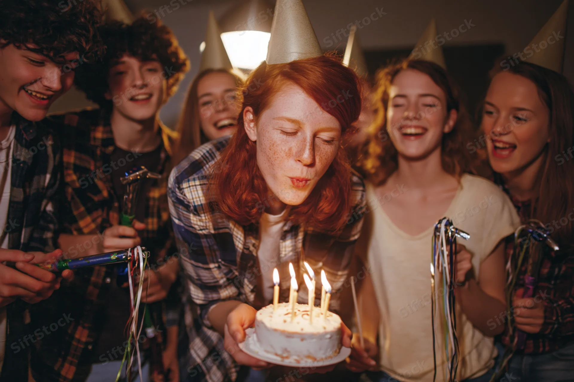 Young woman with red hair blowing out candles on a birthday cake surrounded by friends at a party