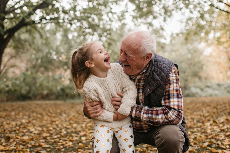 Grandfather hugging laughing granddaughter in autumn park covered in fallen leaves