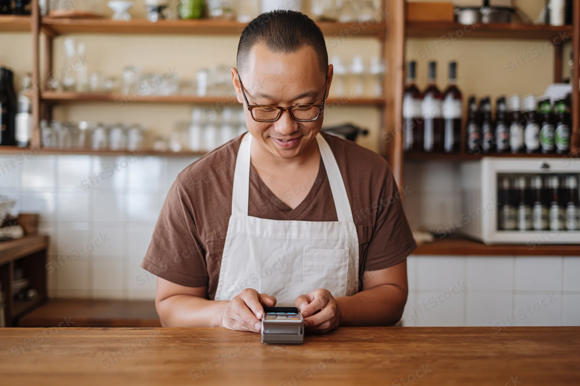 Asian man in apron and glasses using payment terminal at wooden cafe counter