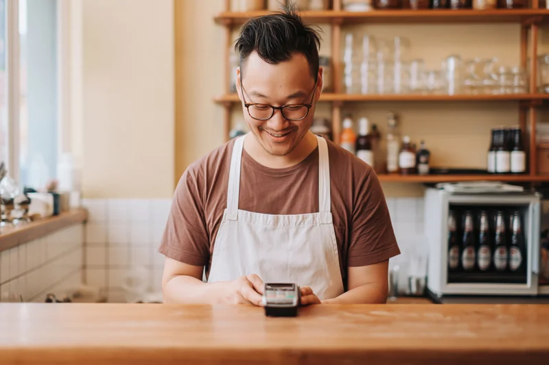 Asian male barista in glasses smiling while processing payment at wooden cafe counter