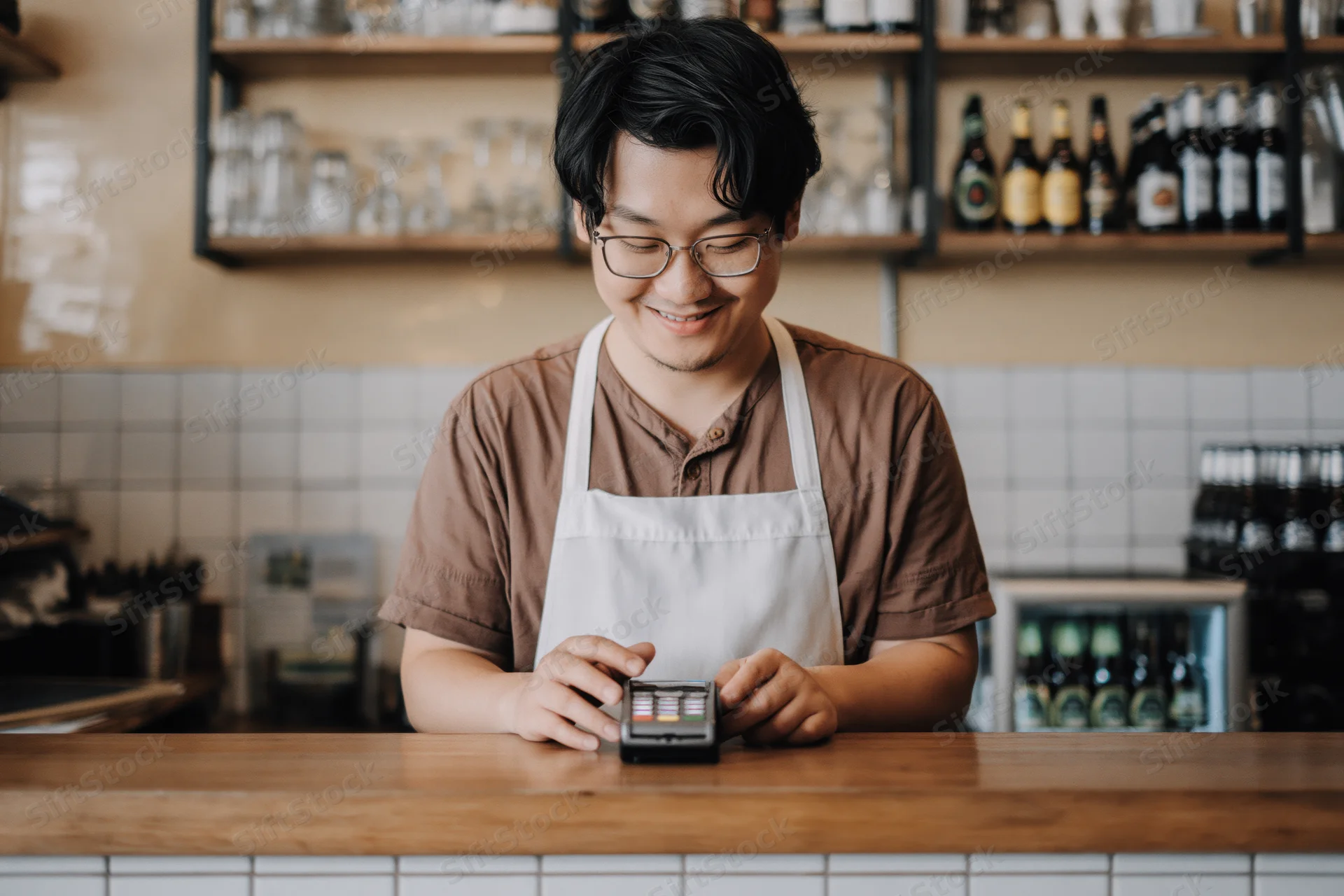 Asian male barista smiling while processing payment on digital terminal behind wooden cafe counter
