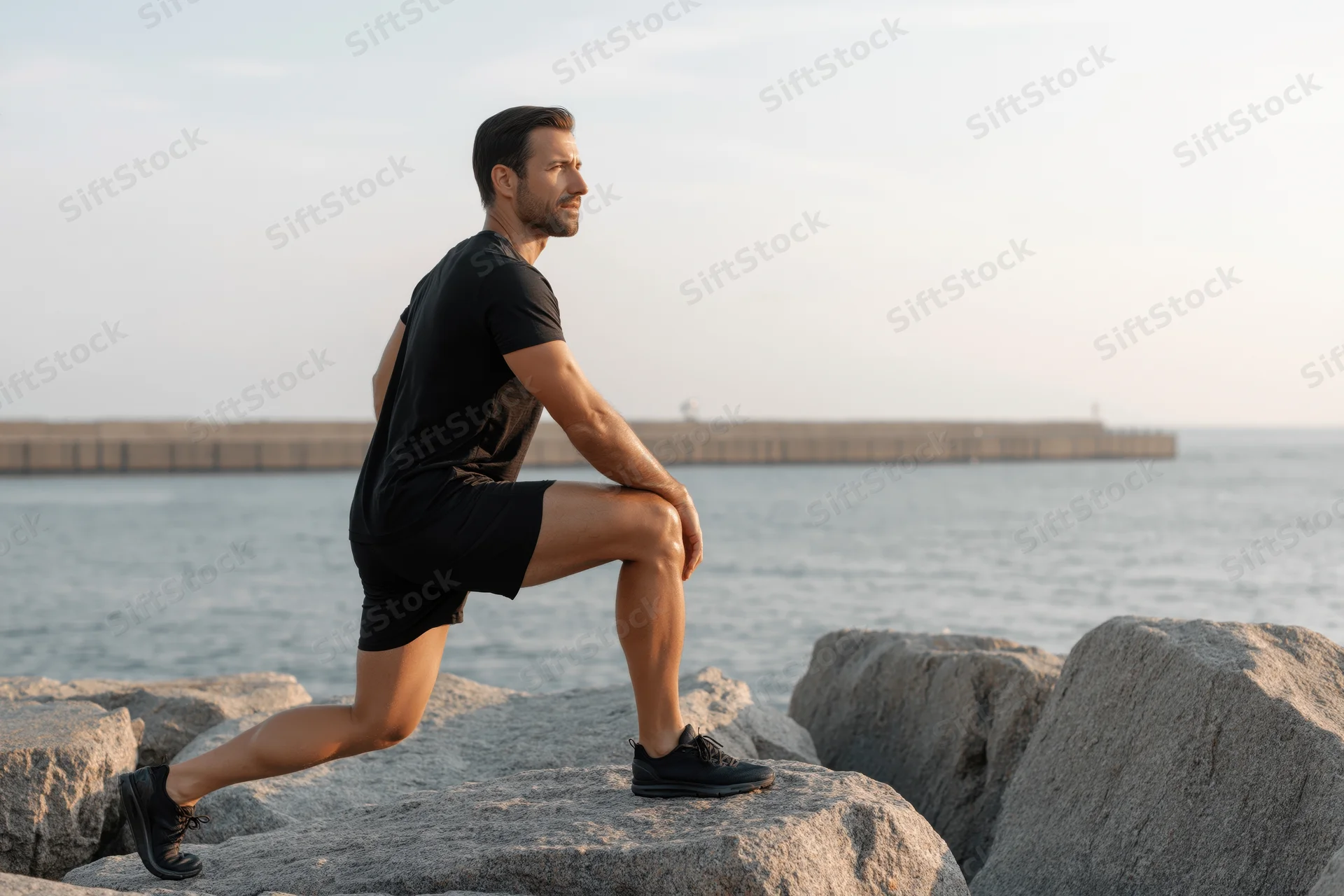 Athletic man performing a deep lunge stretch on coastal rocks by the ocean