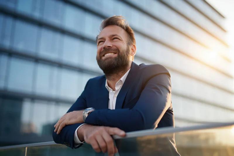 Bearded businessman smiling while leaning on glass railing outside modern office building
