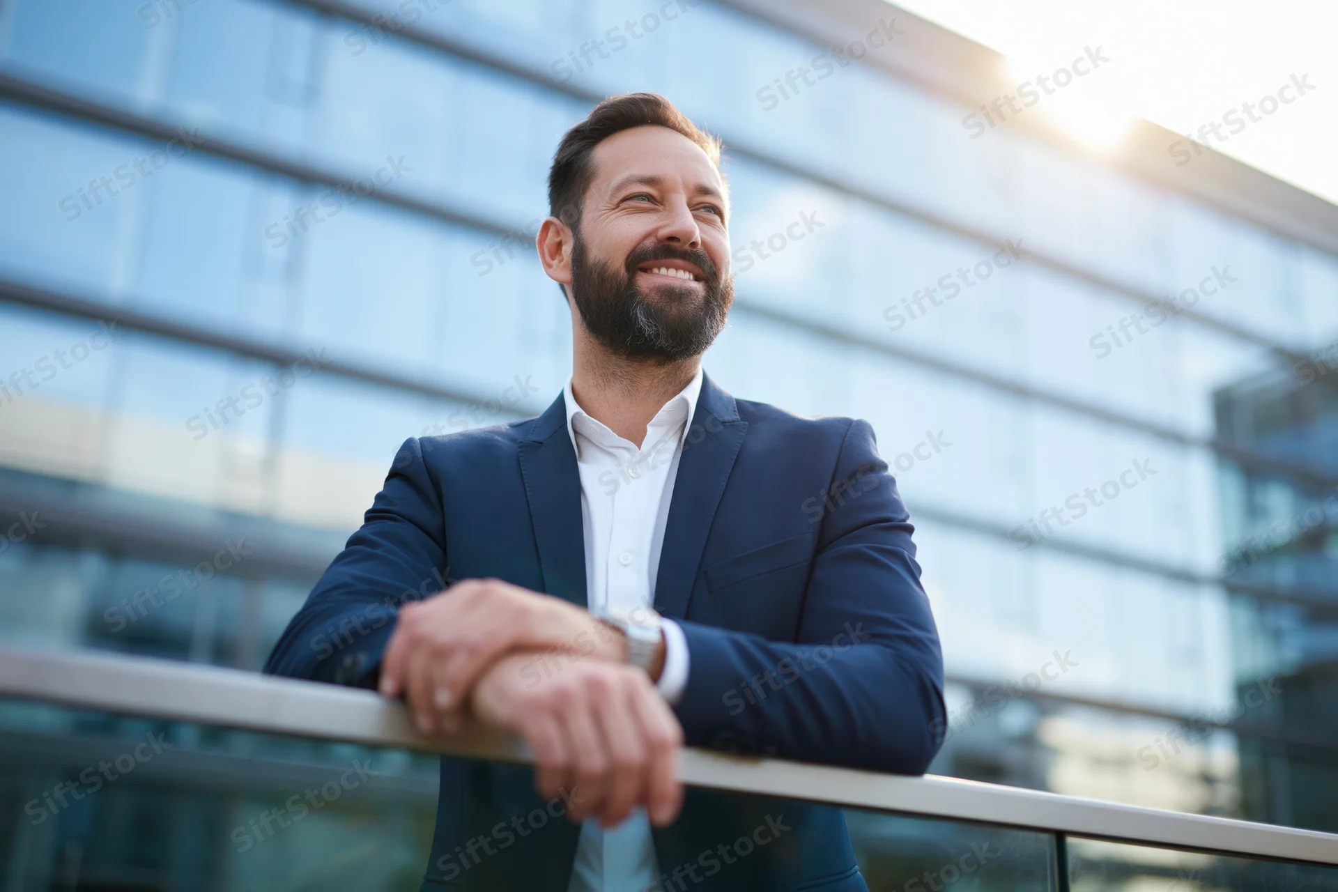 Bearded businessman in suit leaning on glass railing outside modern office building, smiling toward sunlight