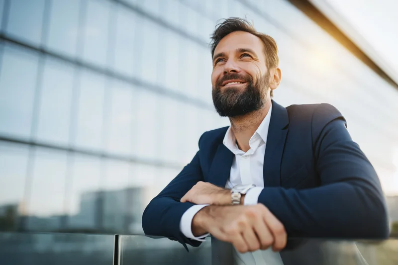 Bearded businessman smiling while leaning on glass railing outside modern office building at sunset