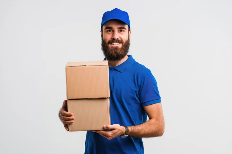 Bearded delivery man in blue uniform smiling while holding stack of cardboard boxes against white background