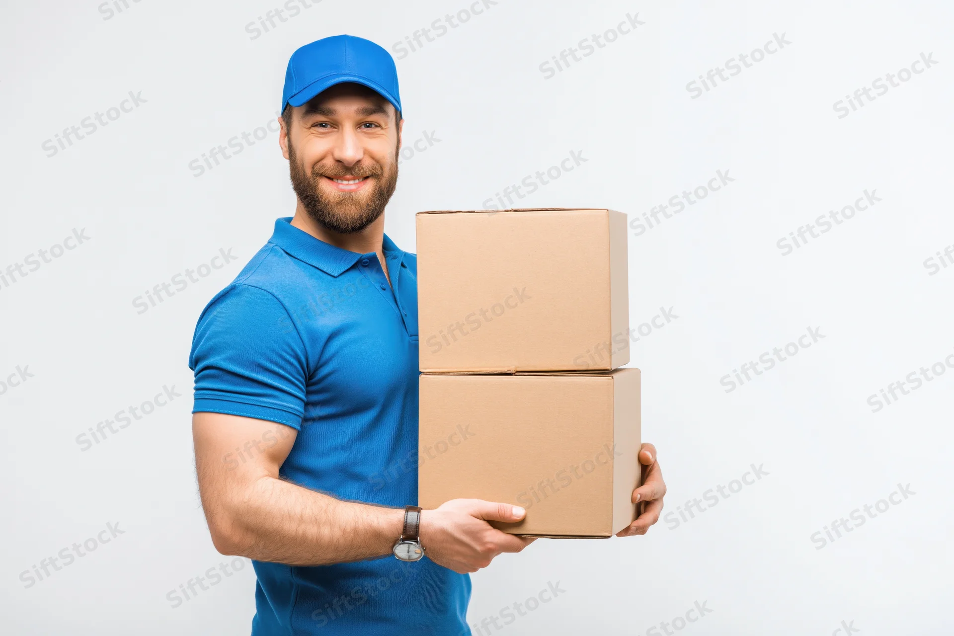 Bearded delivery man in blue uniform smiling while holding stack of cardboard boxes against white background