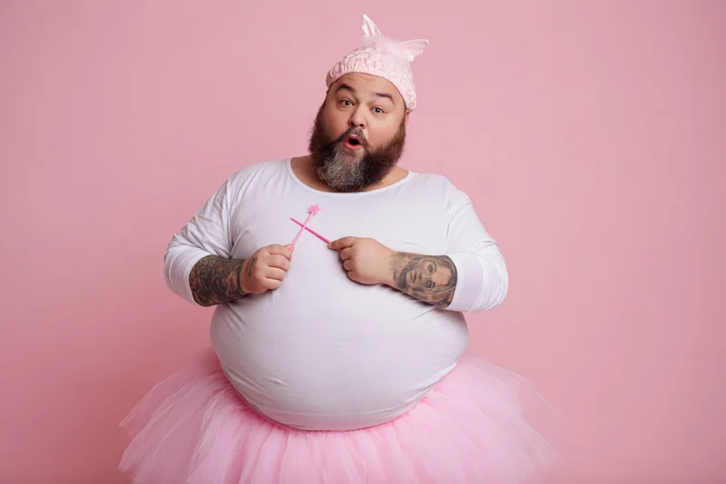 Bearded heavyset man in fairy godmother costume with pink tutu and wand, surprised expression against pink background
