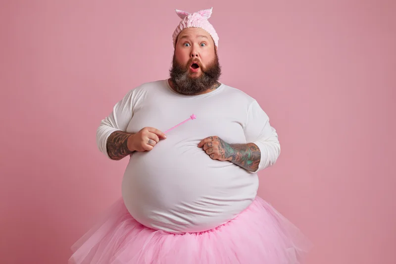 Bearded heavyset man in fairy godmother costume with pink tutu and wand, surprised expression against pink background