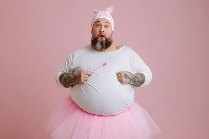 Bearded heavyset man in fairy godmother costume with pink tutu and wand, surprised expression against pink backdrop
