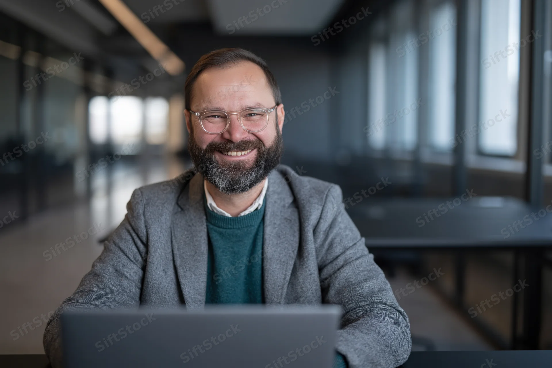 Bearded man smiling at camera while working on laptop in modern office
