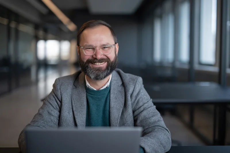 Bearded man smiling at camera while working on laptop in modern office