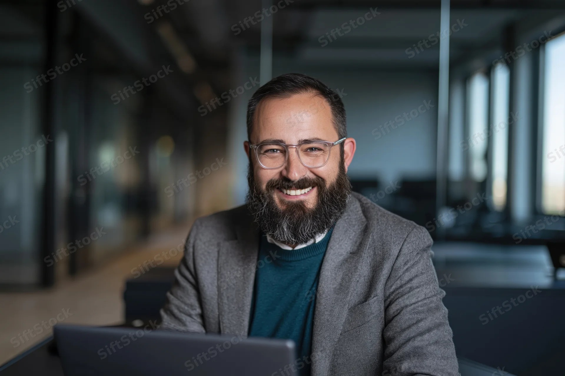 Bearded man in gray suit smiling at camera while working on laptop in modern office