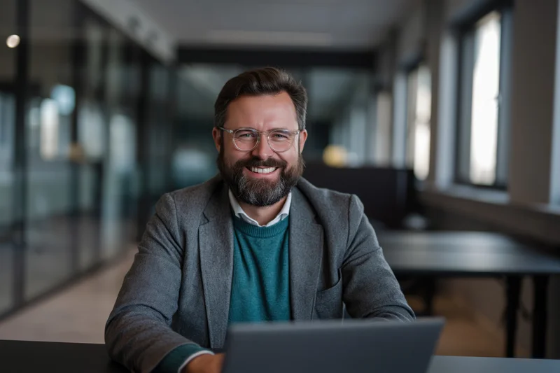 Bearded man smiling at camera while working on laptop in modern office space