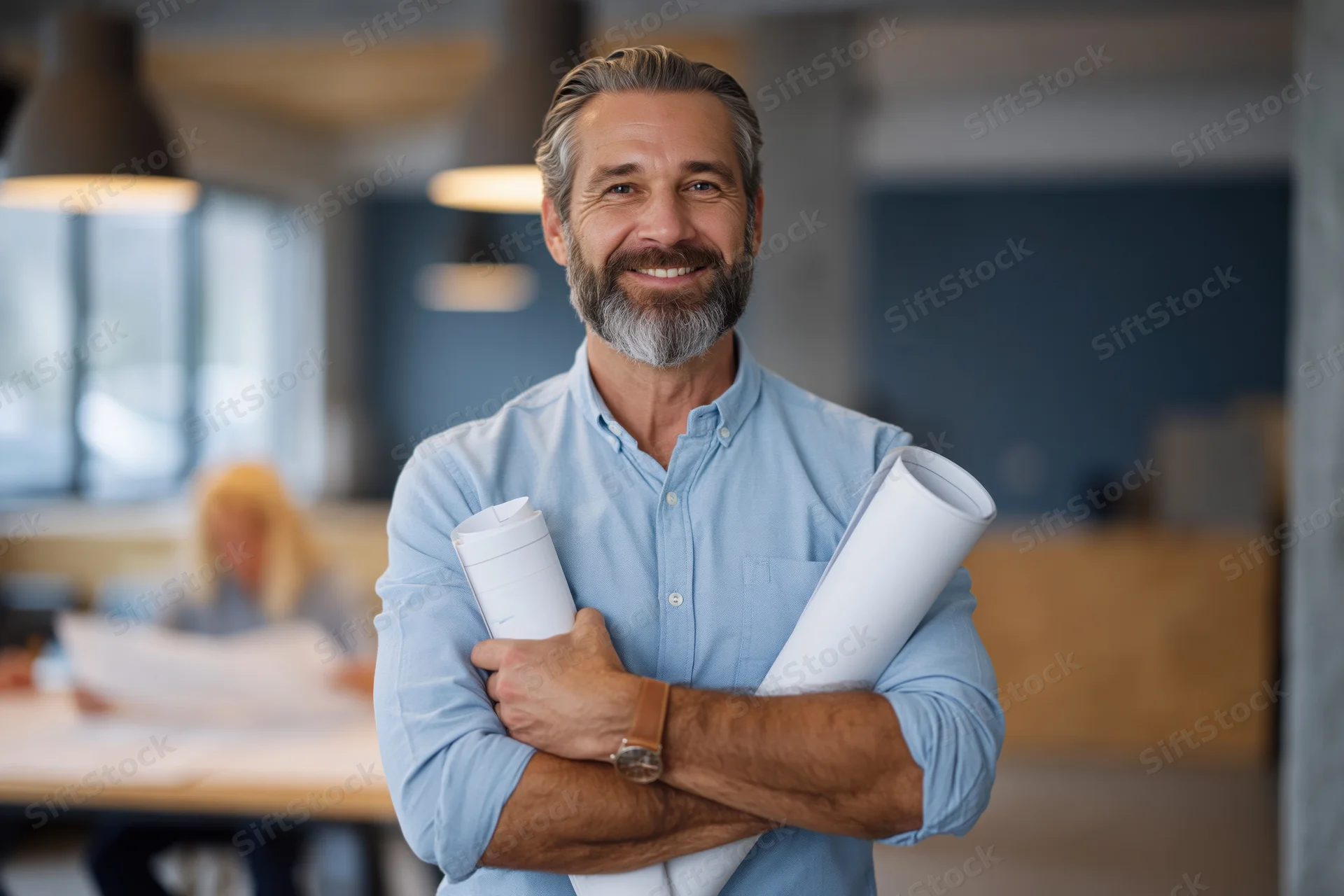 Bearded middle-aged architect smiling with arms crossed holding rolled blueprints in modern office