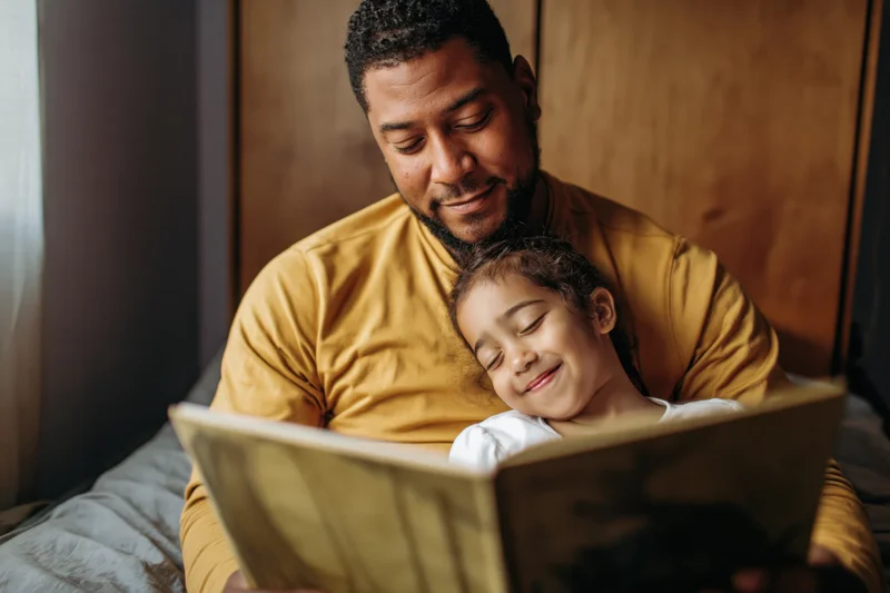 Black father reading a picture book to his young daughter in bed
