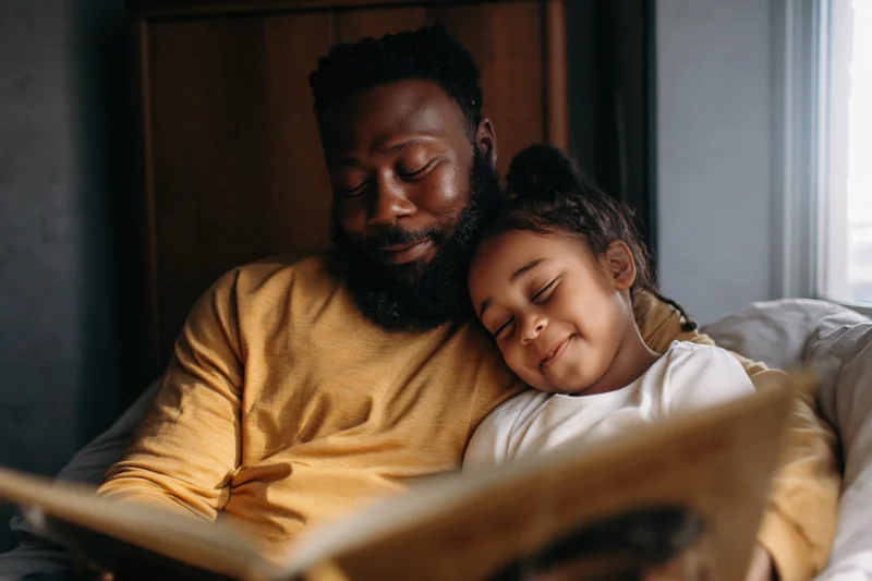 Black father reading a picture book to his young daughter in bed