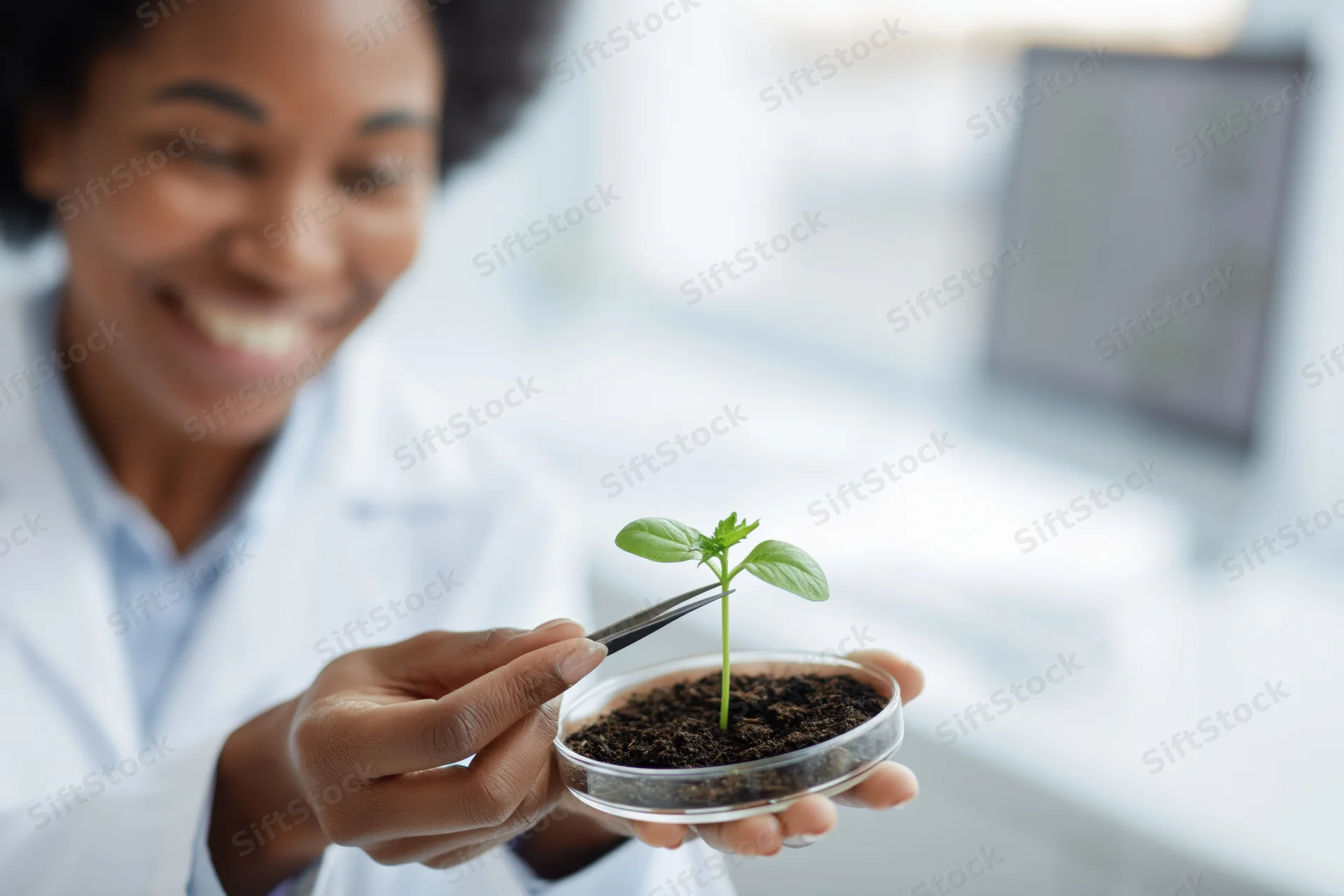 Black female scientist smiling while holding a small plant in a petri dish with tweezers in a lab setting