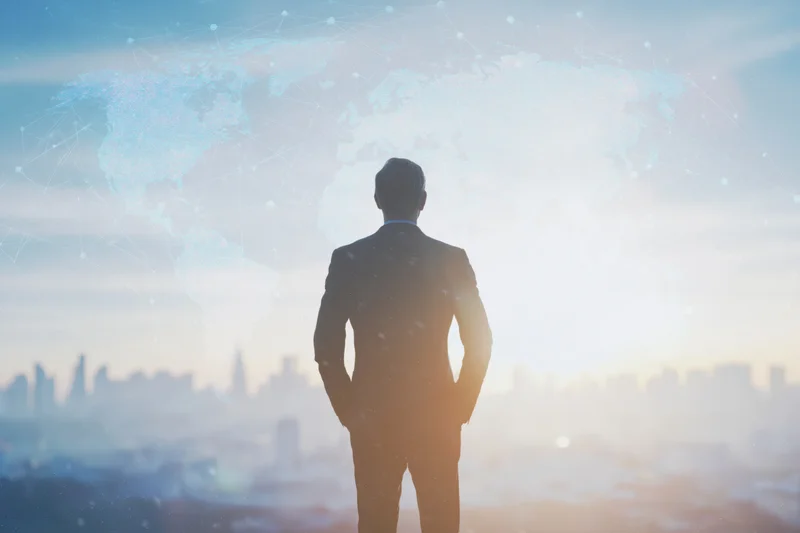 Businessman in dark suit standing with back to camera gazing at city skyline with digital world map overlay