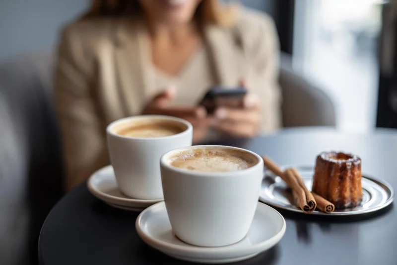 Two white coffee cups on a dark cafe table with a blurred woman using a smartphone in the background