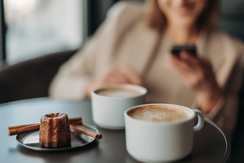 Two white coffee cups and a cinnamon pastry on a dark cafe table with a woman using a smartphone in the background