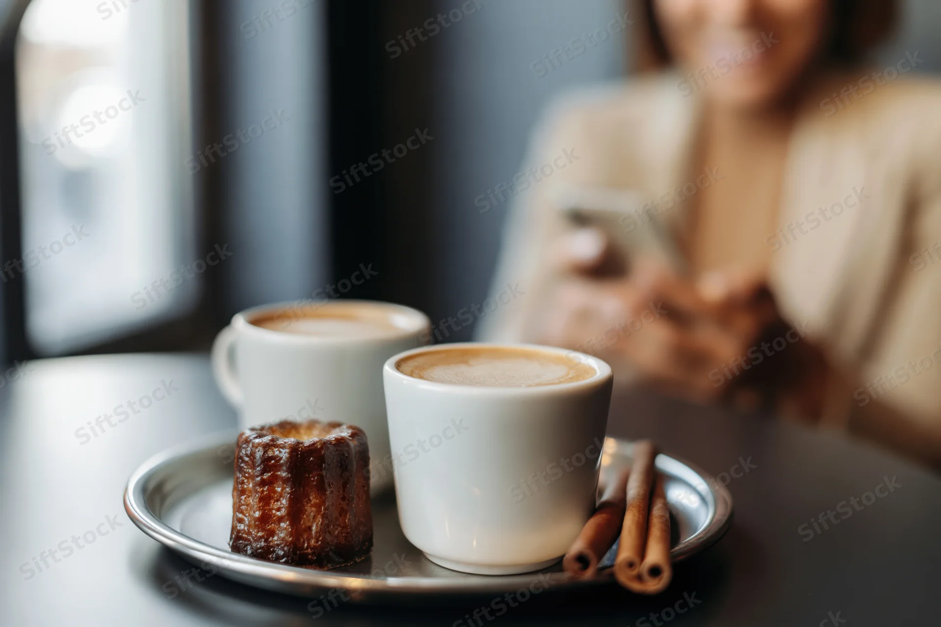 Two white latte cups and a canele pastry on a dark cafe table with a woman using a smartphone in the background