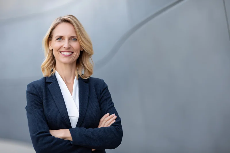 Confident middle-aged businesswoman standing with arms crossed in front of modern gray building exterior