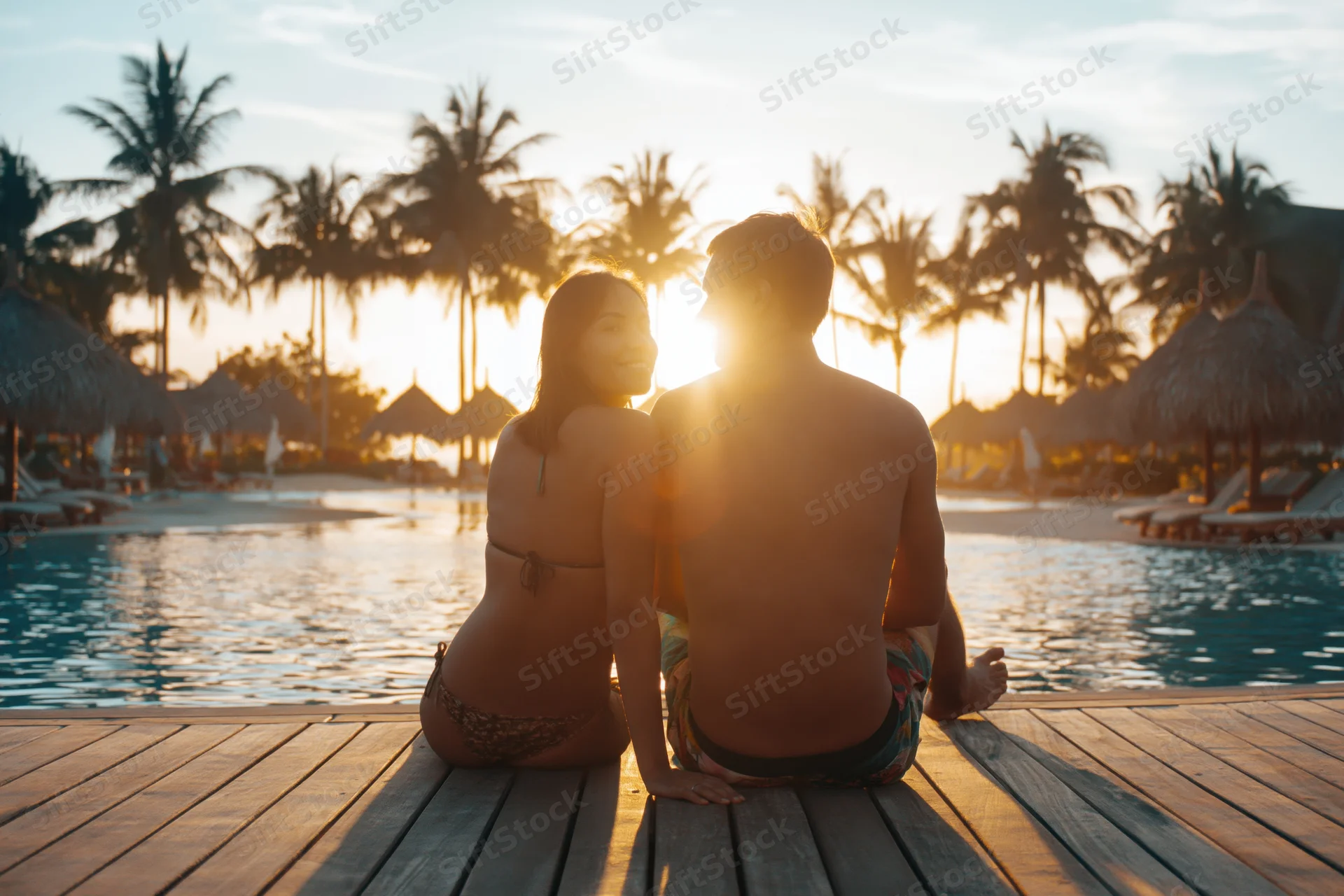 Couple sitting on wooden pool deck facing sunset at tropical resort