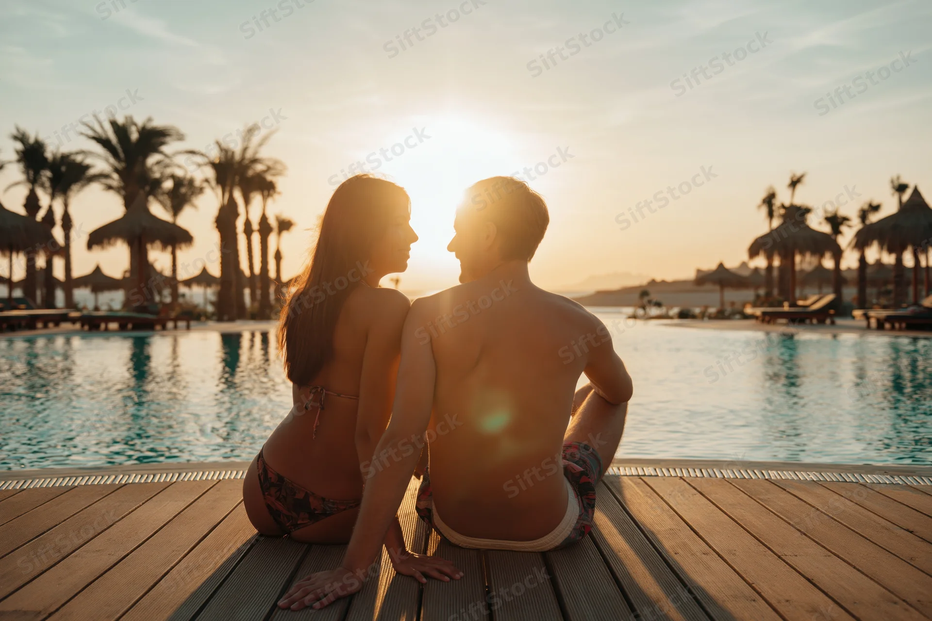 Couple sitting on wooden pool deck facing sunset at tropical resort