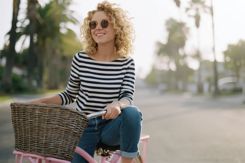 Curly blonde woman smiling while sitting on a pink vintage bicycle with wicker basket on a sunny street