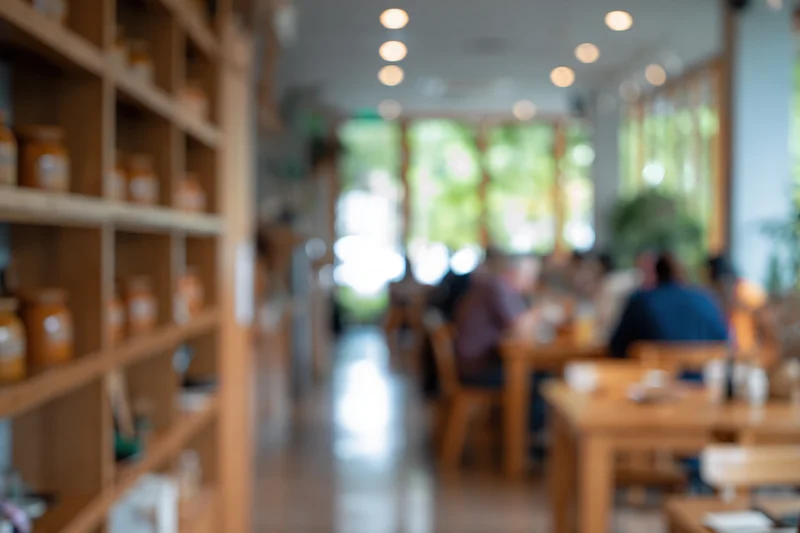 Defocused interior of a casual cafe with wooden shelves and tables, people dining near windows with greenery outside