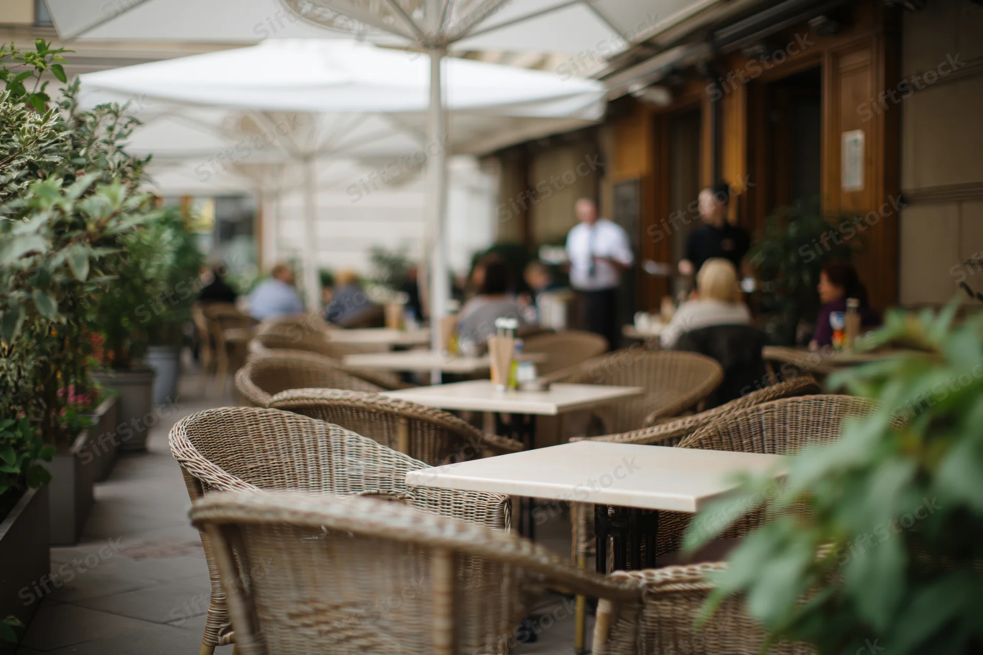 Outdoor cafe terrace with wicker chairs and tables under umbrellas, surrounded by potted plants and greenery, during daylight hours