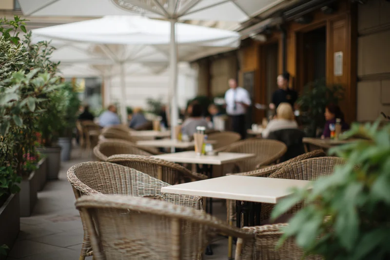 Outdoor cafe terrace with wicker chairs and tables under umbrellas, surrounded by potted plants and greenery, during daylight hours
