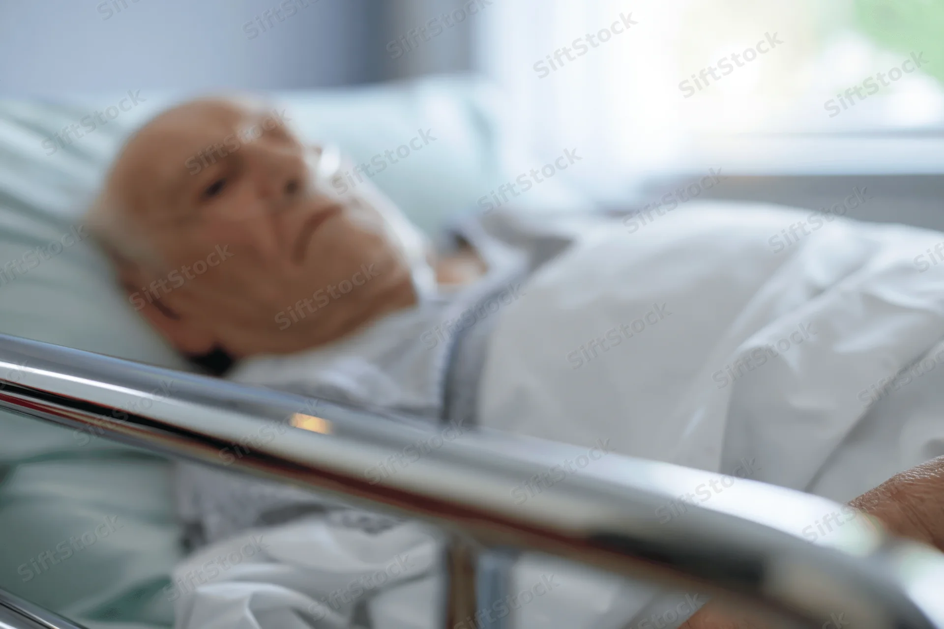 Elderly man lying in hospital bed with oxygen cannula, covered by white blanket, near window