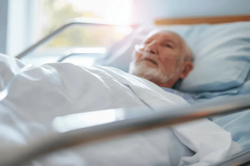 Elderly man lying in hospital bed with oxygen cannula under soft morning light