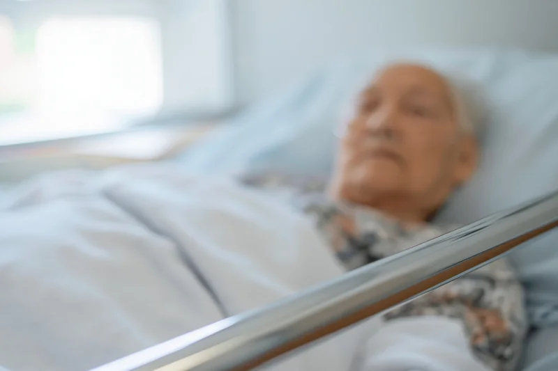Elderly man lying in hospital bed with oxygen cannula, covered by blanket, near window