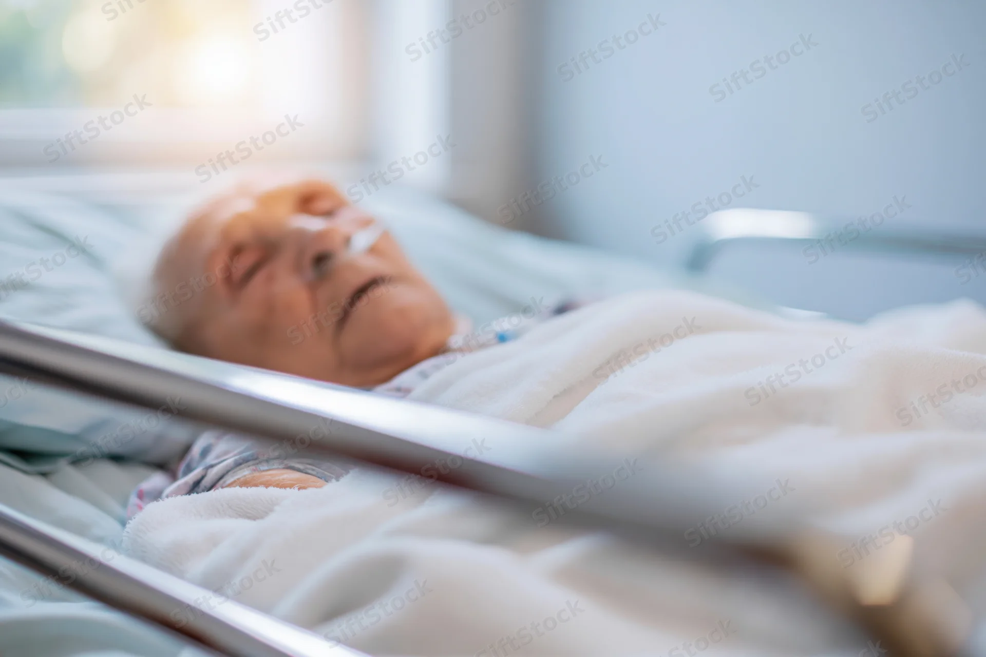 Elderly woman resting in hospital bed with oxygen cannula, covered by blanket near window