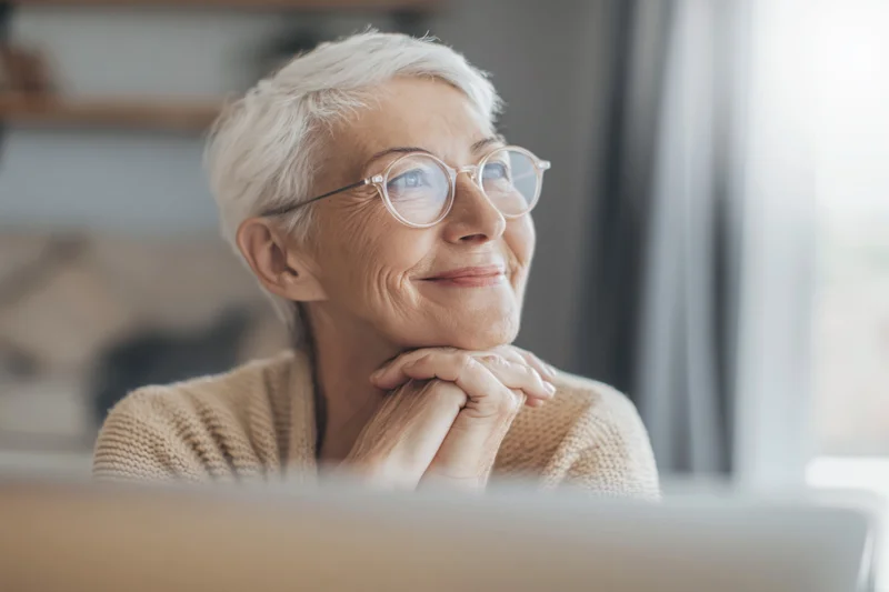 Senior woman with short white hair and round glasses smiling thoughtfully while resting her chin on her hands indoors
