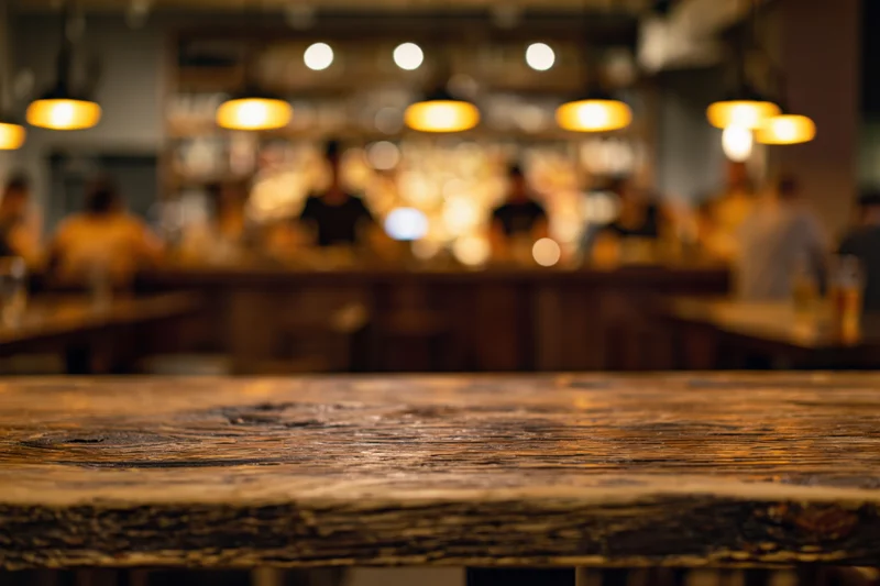 Rustic wooden table surface in foreground with blurred bar interior and warm pendant lights in background