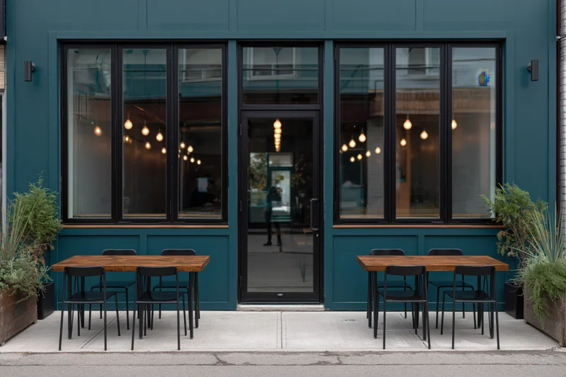 Modern cafe exterior with teal facade, black-framed windows, outdoor wooden tables and chairs on sidewalk