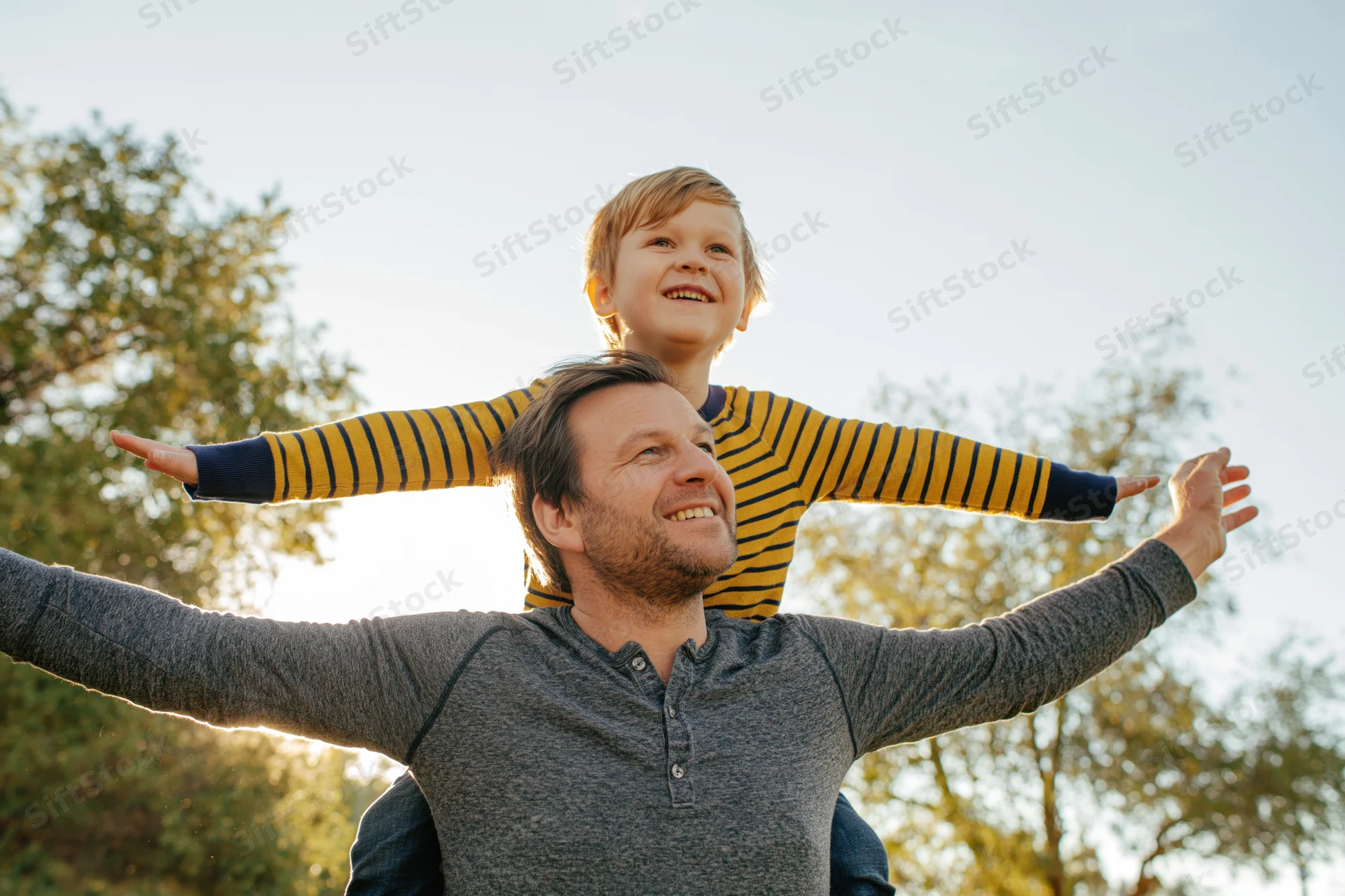 Father carrying young son on his shoulders outdoors with both smiling and arms outstretched