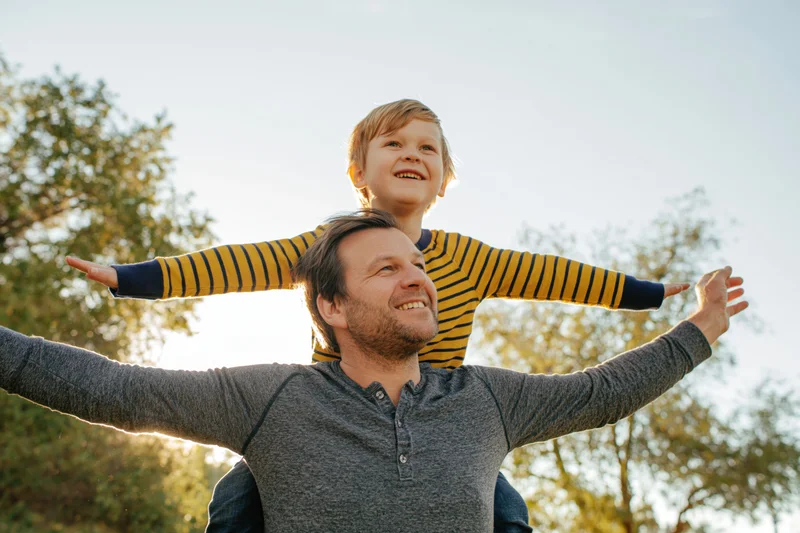Father carrying young son on his shoulders outdoors with both smiling and arms outstretched