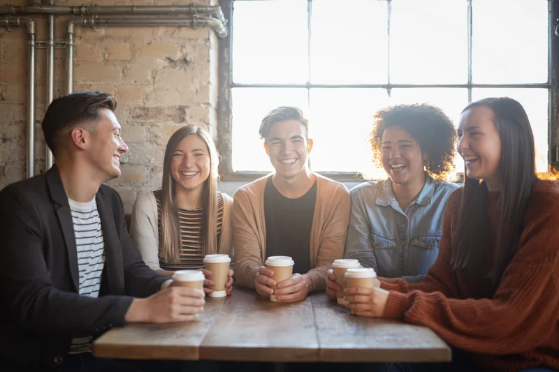 Five young friends laughing together around a wooden table in a cafe