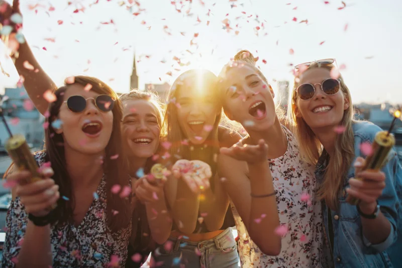 Five young women laughing and celebrating with confetti on a rooftop at sunset