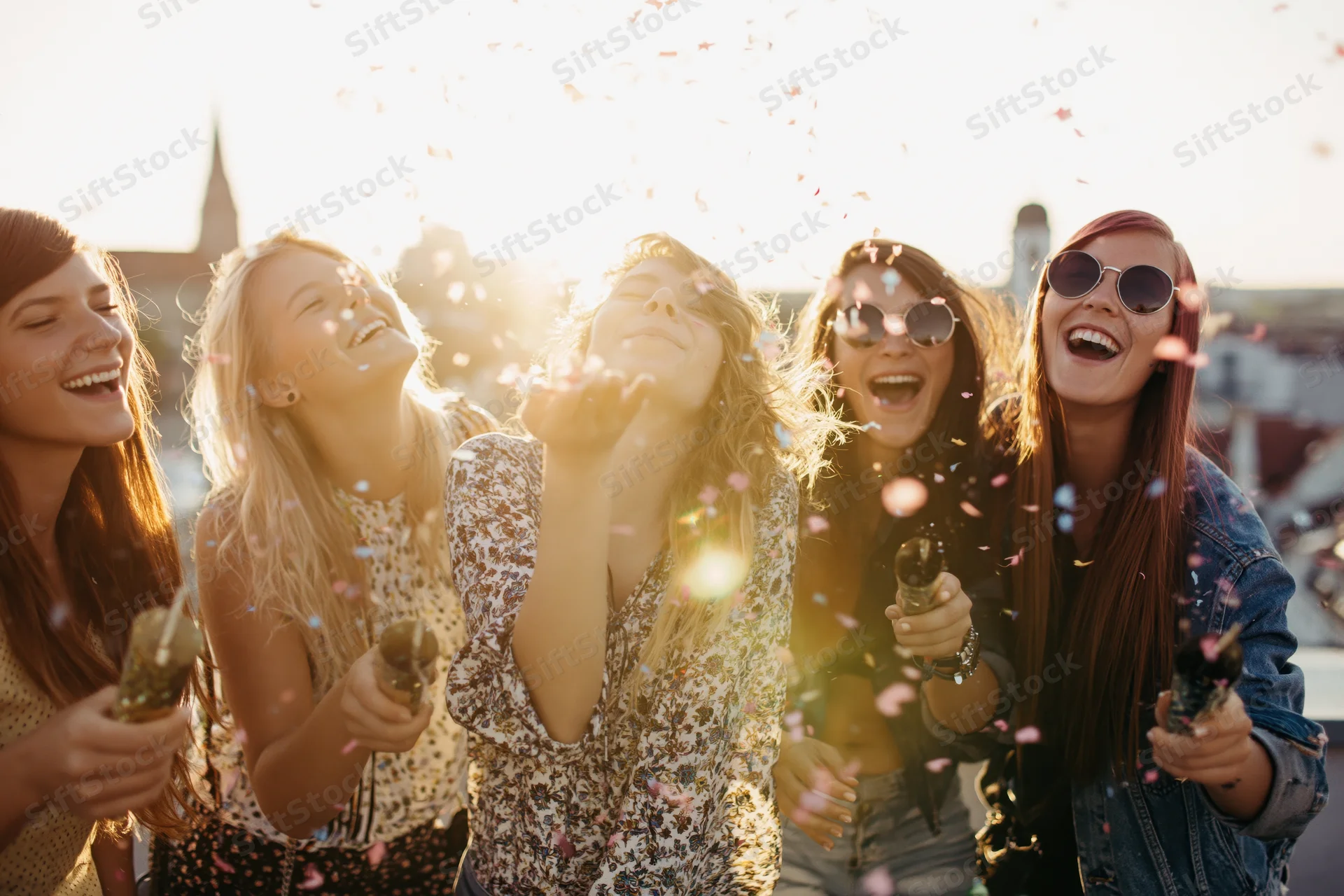 Five young women laughing and throwing confetti on a rooftop at sunset
