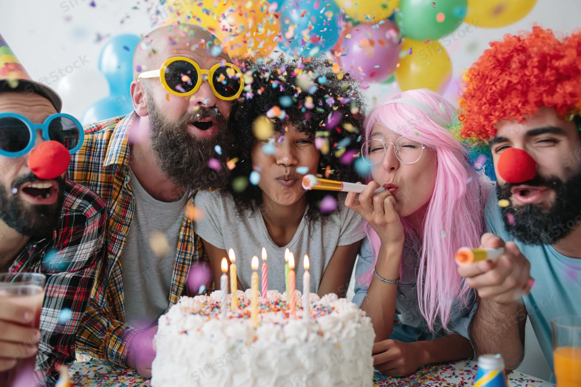 Five friends blowing candles on a birthday cake at a festive party with confetti and balloons