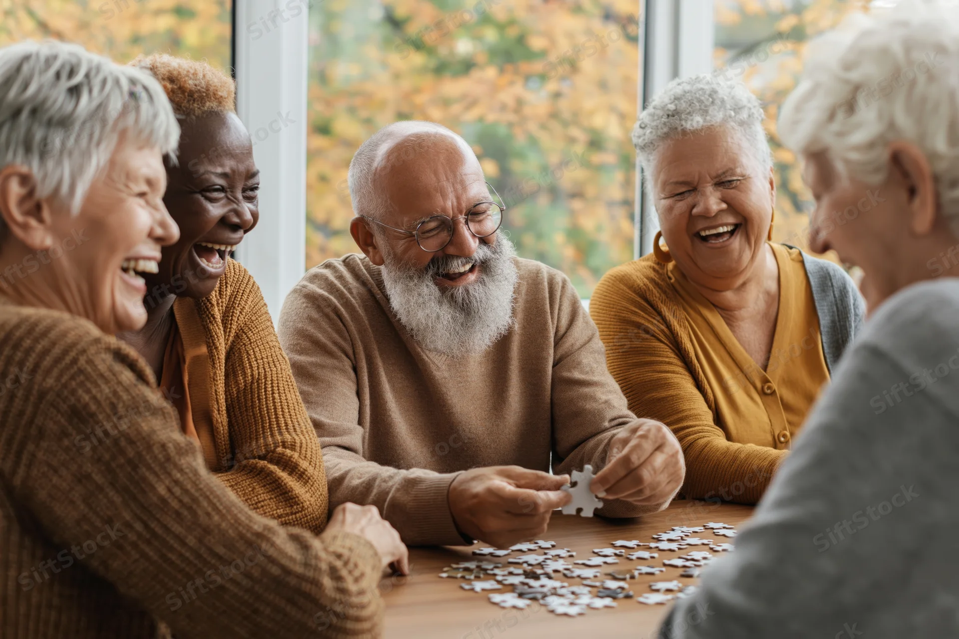 Group of older adults laughing while assembling a puzzle at a table indoors near a window with autumn foliage outside