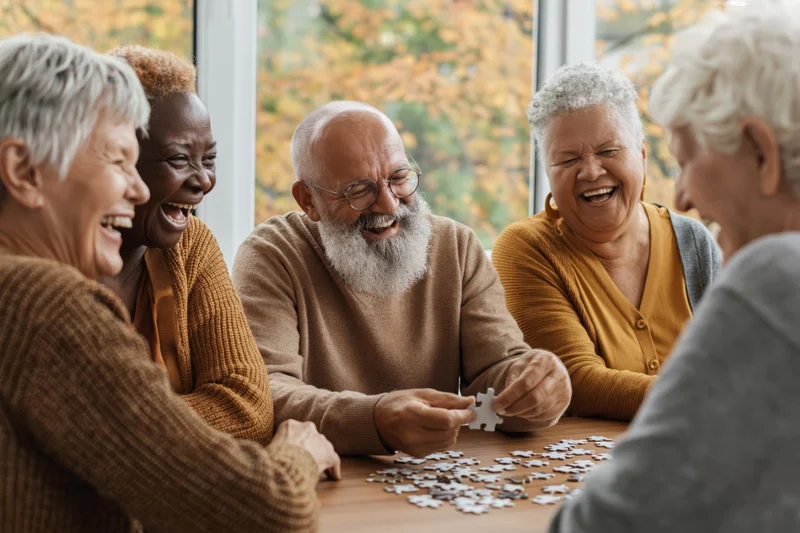 Group of older adults laughing while assembling a puzzle at a table indoors near a window with autumn foliage outside