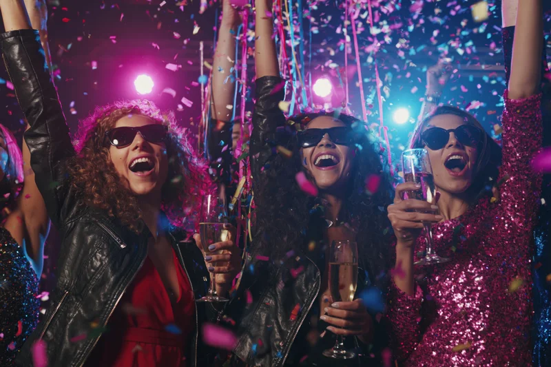 Three women cheering with arms raised and champagne glasses at a nightclub party with confetti falling