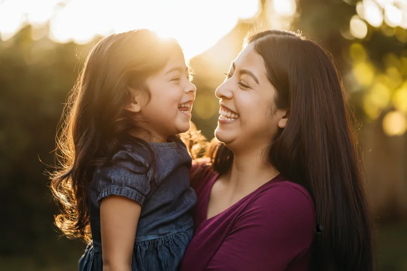 Latina mother holding her young daughter in her arms both laughing outdoors at golden hour